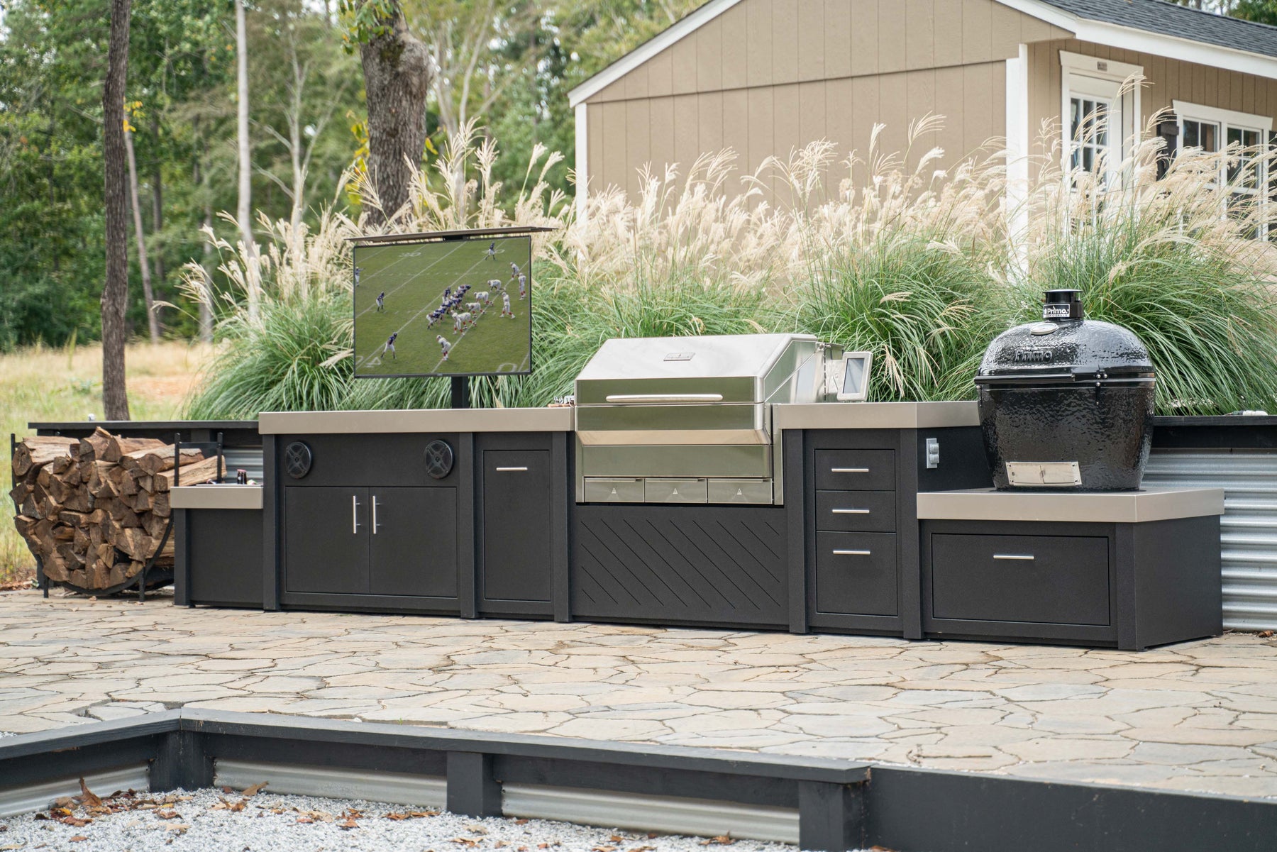 Stono Outdoor Living kitchen island, built-in gas grill, a Primo kamado grill, an outdoor refrigerator, an outdoor TV, a firewood rack, and a stainless steel sink with faucet.